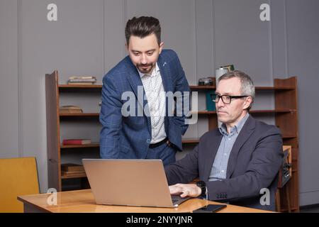 Mature businessman in suit and his pensive younger colleague are working on the laptop in the office Stock Photo