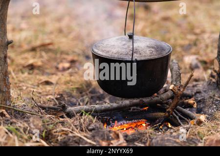 Black large tourist pot hangs over burning flame of campfire in the forest.  Cooking dinner on