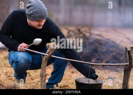 The guy holds a large spoon in one hand, and with the other he lifts the lid of the cauldron hanging over the fire. Stock Photo