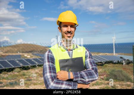 A smiling engineer with folded arms at a solar farm holding a folder. Stock Photo