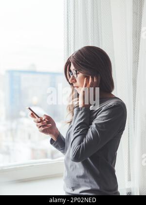 Thoughtful woman with eyeglasses looks at her smartphone. Information in online media. Wireless headphones under hair. Stock Photo