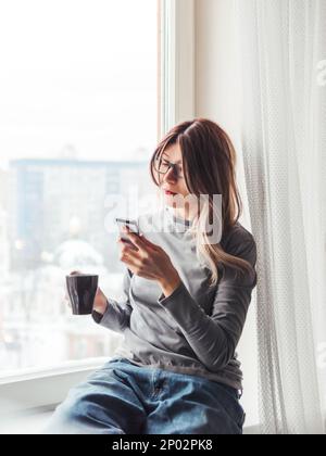 Thoughtful woman with eyeglasses and cup of hot coffee looks at her smartphone. Information in online media. Stock Photo