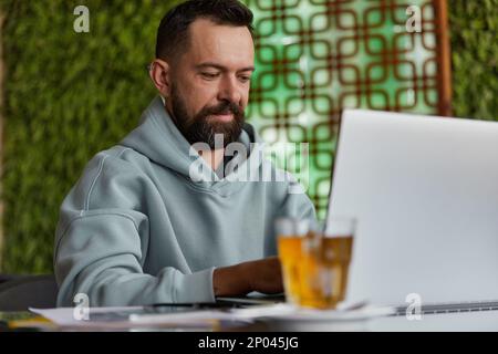 Smiling bearded man in casual clothes in cafe using laptop Stock Photo