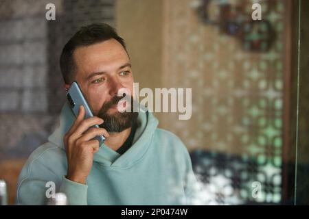 Young businessman talking on his phone indoor through glass. Bearded man in casual clothes using his smartphone. Stock Photo