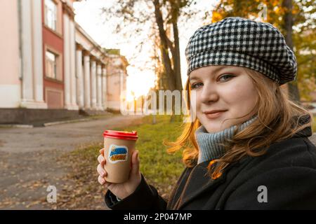 Ukraine. Sunset. City Park. Girl sits on a bench and holds cup of coffee with patriotic sticker "Glory to Ukraine" in her hands. Walks in open air Stock Photo