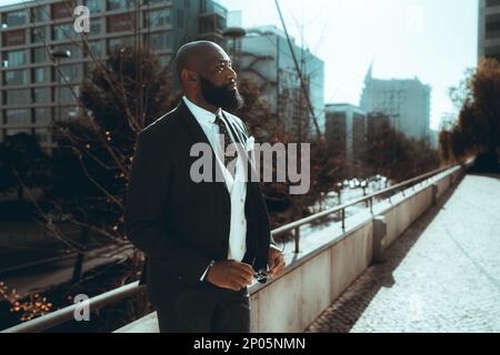 A bald, well-dressed black man leans against an iron railing on the sidewalk, clutching a pair of sunglasses; dressed to impress in a sharp suit and t Stock Photo
