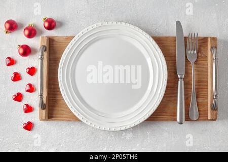 An empty white ceramic plate with cutlery on a wooden tray surrounded by red christmas balls and hearts on a light concrete table. Food display mockup Stock Photo