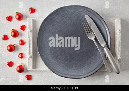 An empty gray ceramic plate with cutlery on a white wooden tray surrounded by red christmas balls and hearts on a light concrete table. Food display m Stock Photo
