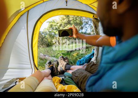 View from in tent of diverse couple camping, lying in tent taking selfie in countryside Stock Photo