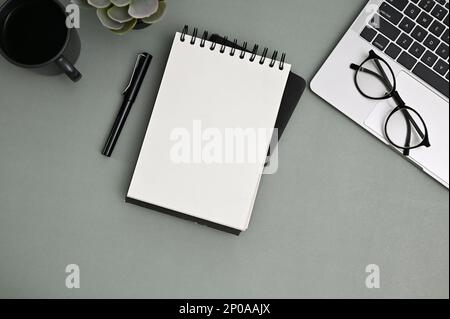 Modern workspace top view with blank empty page notepad, pen, eyeglasses, laptop, coffee cup and decor on grey background. Stock Photo