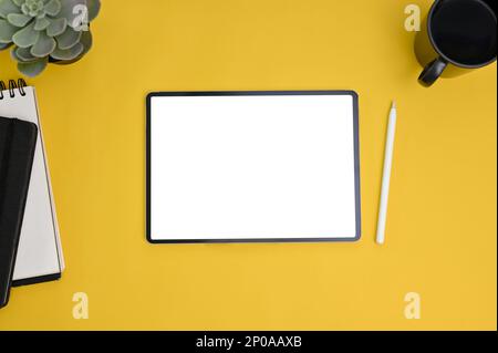 Workspace top view with digital tablet white screen mockup, stylus pen, notebook, coffee cup and decor plant on yellow background. Stock Photo