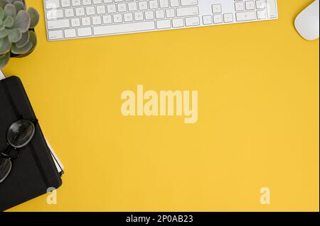 Top view workspace composition with pc keyboard, wireless mouse, eyeglasses, notebook, decor plant and copy space on yellow background. Stock Photo