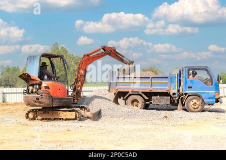 Mini excuvator loading gravel to dumper truck in construction site. Stock Photo