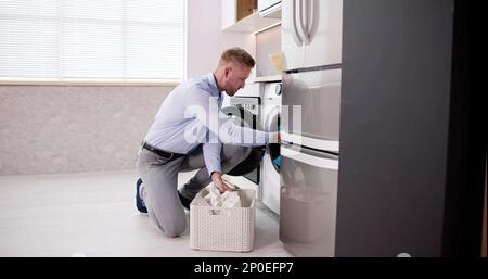 Young Man Loading Clothes Into Washing Machine In Kitchen Stock Photo