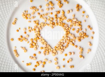 Yellow dried peas in a shape of heart spilled on a white plate. Raw groats food ingredient background. Love for cooking concept Stock Photo