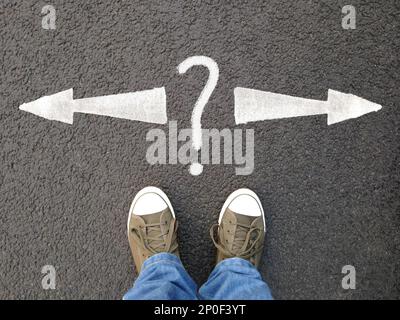 feet in canvas shoes standing on asphalt from personal perspective, road markings with arrows pointing left and right with question mark Stock Photo