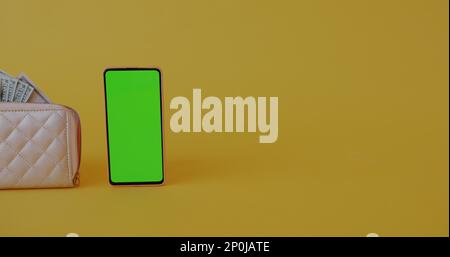 Green screen chroma key smartphone and purse full of cash on yellow background with no people visible. Mobile application and finance concept. Stock Photo