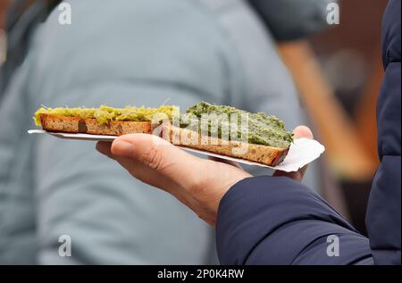 A visitor of Prague farmers market holds a paper tray with a snack, toast with herb pesto. Stock Photo