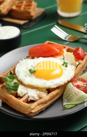 Delicious Belgian waffle with fried egg, salmon, cream cheese and vegetables served on green table, closeup Stock Photo