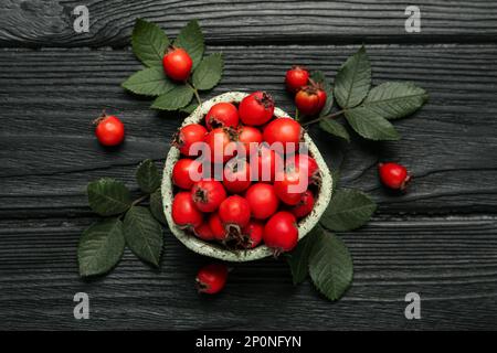 Ripe rose hip berries with green leaves on black wooden table, flat lay Stock Photo