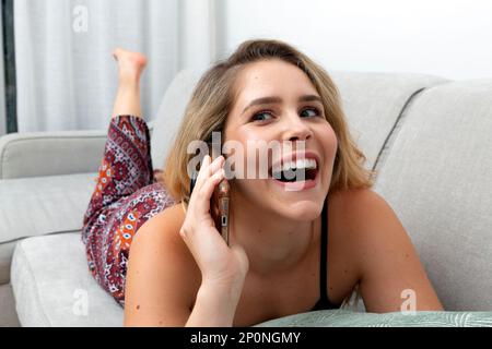 The young blonde woman is having a friendly conversation on the phone while lounging on the couch with a smile on her face. Stock Photo