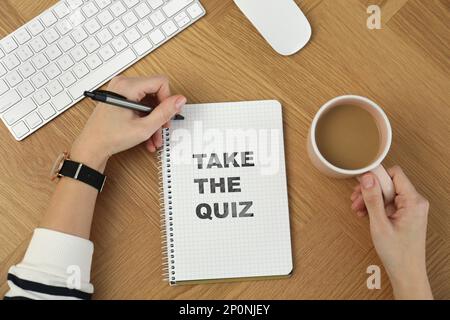 Woman with cup of coffee writing phrase Take the quiz in notebook at wooden table, top view Stock Photo