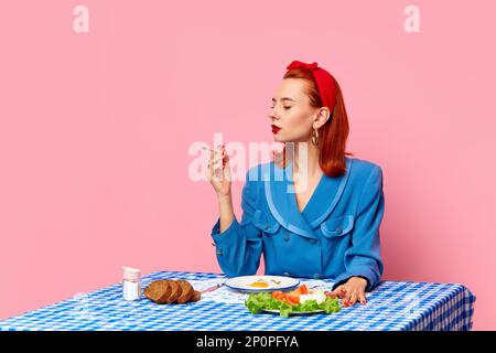 Stylish redhead woman sitting at table, smoking and having english breakfast with fried eggs against pink studio background. Food pop art photography. Stock Photo