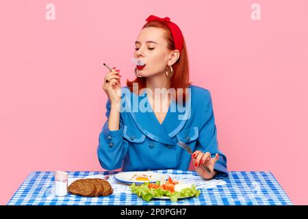 Stylish redhead woman sitting at table, smoking and having breakfast with fried eggs and salad against pink studio background. Food pop art Stock Photo