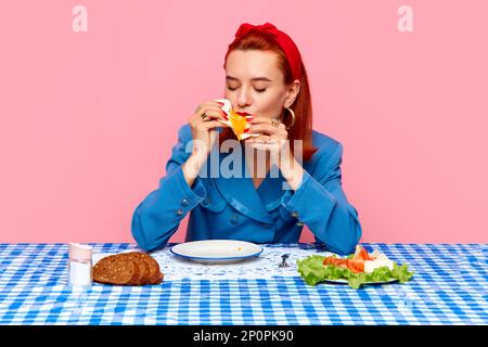 Young redhead woman sitting at table with checkered tablecloth and eating breakfast with fried eggs against pink studio background. Food pop art Stock Photo