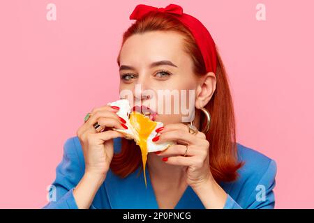Close-up portrait of young redhead woman eating fried eggs against pink studio background. Liquid yolk. Food pop art photography. Stock Photo