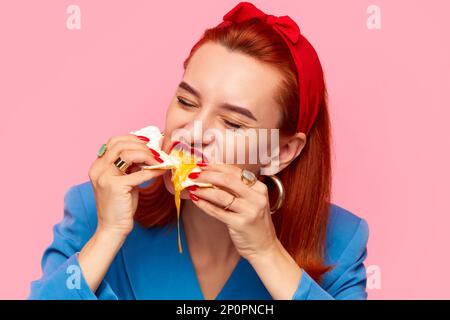 Close-up portrait of young redhead woman eating fried eggs against pink studio background. Liquid yolk. Food pop art photography. Stock Photo