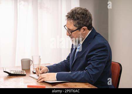 Businessman in his 50s with beard and dressed in a suit and tie sitting at his office desk writing with a pencil on a notepad Stock Photo