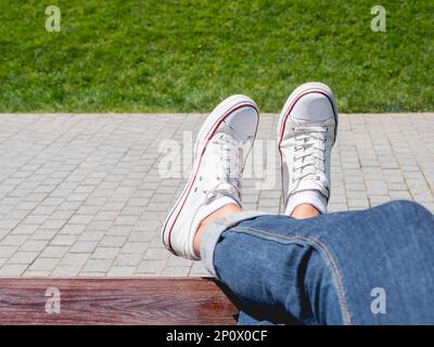 Woman feet in white sneakers on grass lawn background. Female has a rest on wooden bench in urban park at sunny day. Summer vibes. Slow living. Stock Photo
