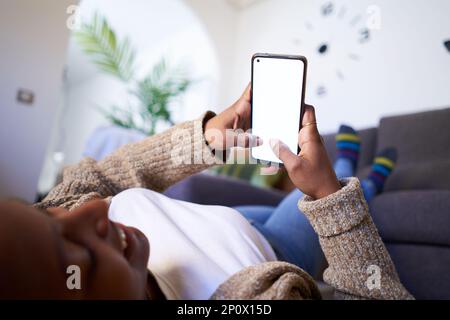 Young black lady using phone while lying and relaxing on the floor putting feet on couch at home. Stock Photo