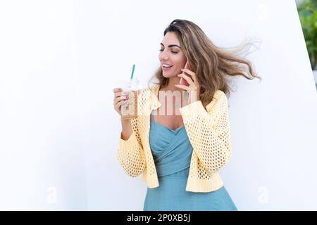 Portrait of beautiful woman in green summer dress on white background natural daylight, holding iced coffee cappuccino talking on phone smiling Stock Photo