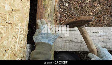 hands of a carpenter in gray fabric gloves hold a long nail and a hammer over old boards and a log, fixing the details of a wooden structure Stock Photo