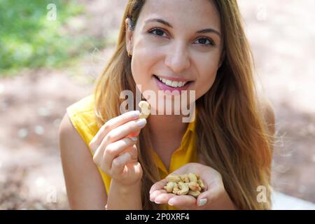 Natural healthy girl picking cashew nuts from her hand in the park. Looks st camera. Stock Photo