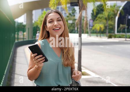 Pretty young Brazilian brown-haired woman looking at camera using smartphone outdoors. Long-haired lady wears elegant tank top. Concept of city life, Stock Photo