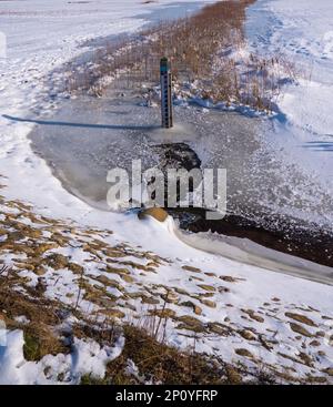 Winter landscape of a partially frozen ditch, withe a dipstick in the middle. And reeds on the background. Stock Photo