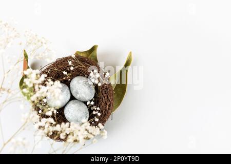 Brown nest of twigs with three gray Easter eggs, feathers, dried magnolia leaves and sprigs of gypsophila on gray background. Minimalistic Easter conc Stock Photo