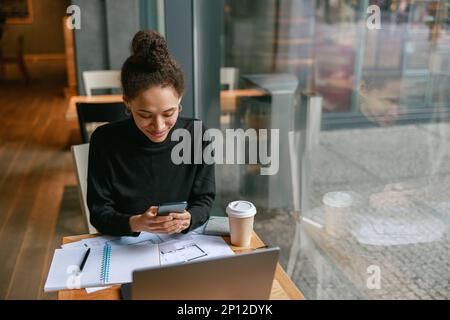 Woman architect use phone during working in cafe sitting near window. Distance work concept Stock Photo