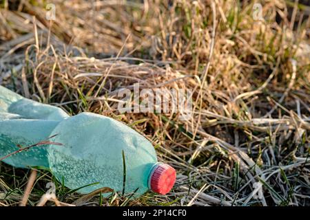 Crumpled plastic bottle covered with ice and snow in frozen dry grass. Green with red bottle cap. Plastic waste left in nature. Stock Photo