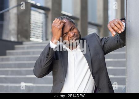 Senior African American businessman suffering from severe headache, pressure. He is standing outside the office center, leaning against the wall, holding his head, grimacing in pain. Stock Photo