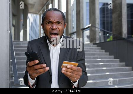 Worried shocked senior african american businessman in suit standing outside near office center. Holds a blocked credit card and phone. Bankruptcy, fraud. Screams angrily at the camera. Stock Photo