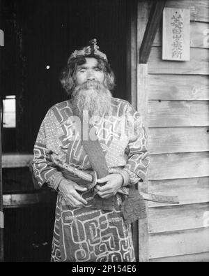 Ainu chief wearing a headdress and holding a sword standing in a doorway, 1908. Stock Photo