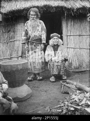 Ainu man and seated woman at the entrance of a hut, 1908. Stock Photo