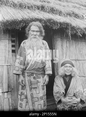 Ainu man and seated woman at the entrance of a hut, 1908. Stock Photo