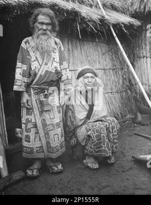 Ainu man and seated woman at the entrance of a hut, 1908. Stock Photo