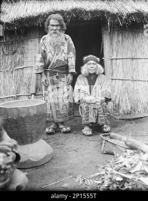 Ainu man and seated woman at the entrance of a hut, 1908. Stock Photo