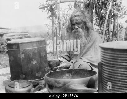 Ainu man seated outdoors on a mat covered with clay containers, 1908. Stock Photo